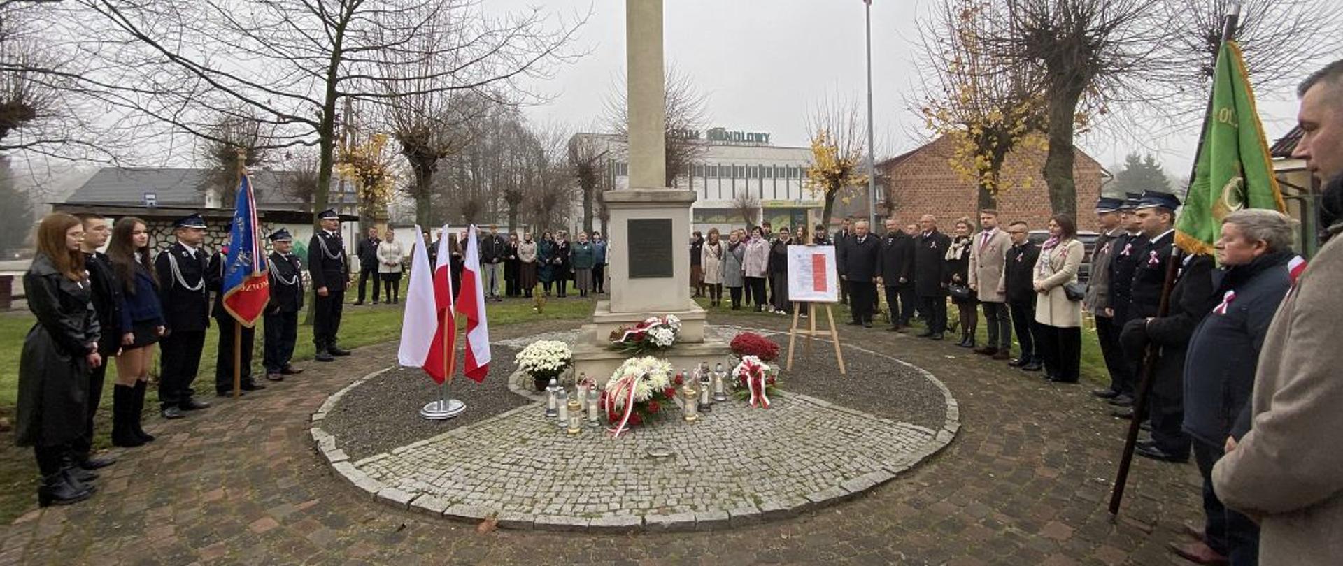 Szeroki plan przedstawia uroczystość patriotyczną z okazji Narodowego Święta Niepodległości w pochmurny, jesienny dzień. Centralnie usytuowany jest pomnik-kolumna z orłem na szczycie. U podnóża pomnika, na okrągłym brukowanym placu, leżą wieńce i kwiaty w barwach narodowych, a także palą się znicze. Wokół pomnika stoi duża grupa ludzi – po lewej młodzież i osoby w mundurach, po prawej cywile i kolejni mundurowi, wszyscy w postawie uroczystej. W tle widać maszt z polską flagą oraz bezlistne drzewa i zabudowania.