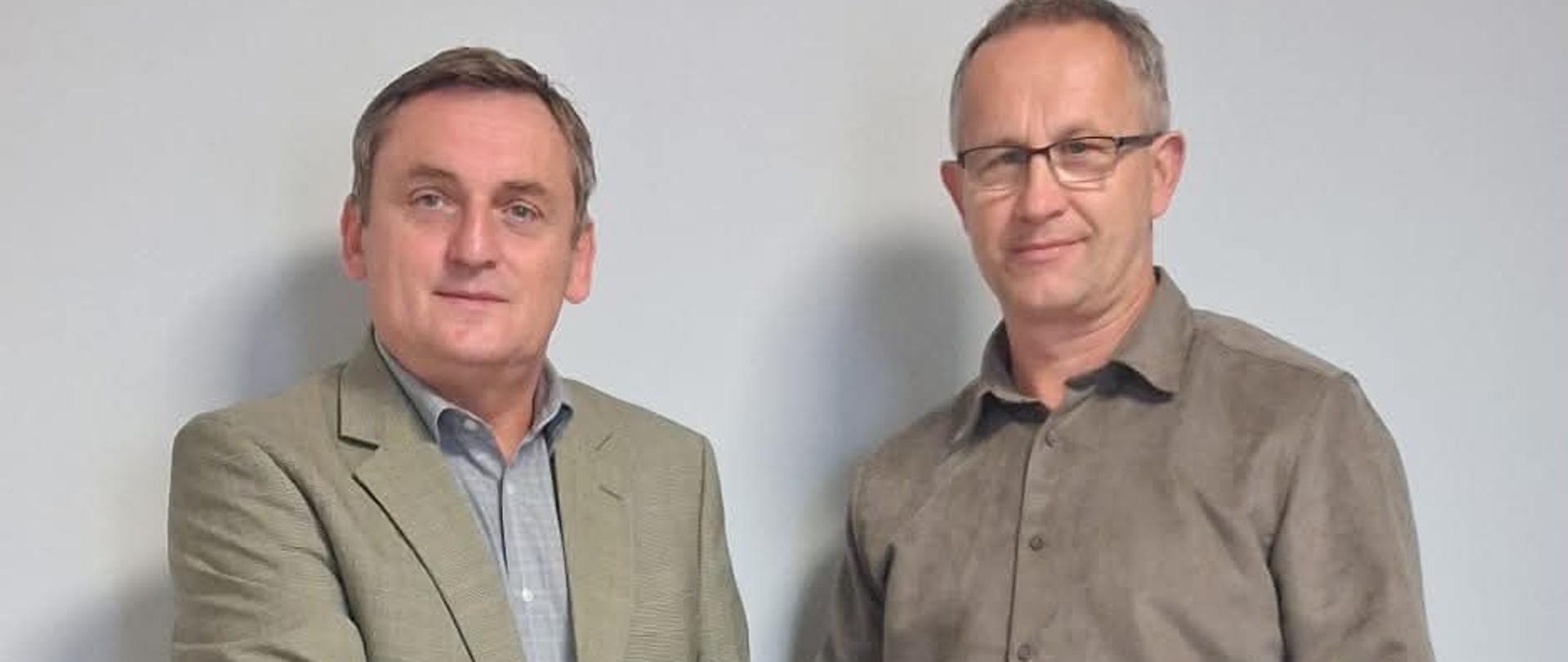 Dwóch mężczyzn w średnim wieku stoi na tle ściany, ściskają się za ręce. drugi mężczyzna od lewej strony trzyma w ręce dodatkowo granatową teczkę z napisem Gmina Wietrzychowice.