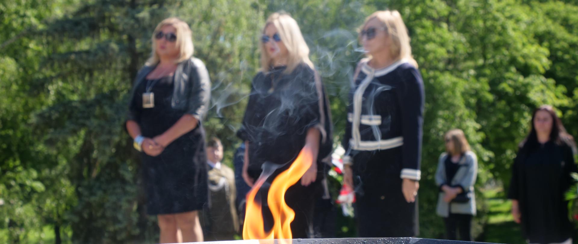 Zdjęcie przedstawia odpalony, duży znicz na pierwszym planie. W tle stoją trzy kobiety w czarnych ubraniach
