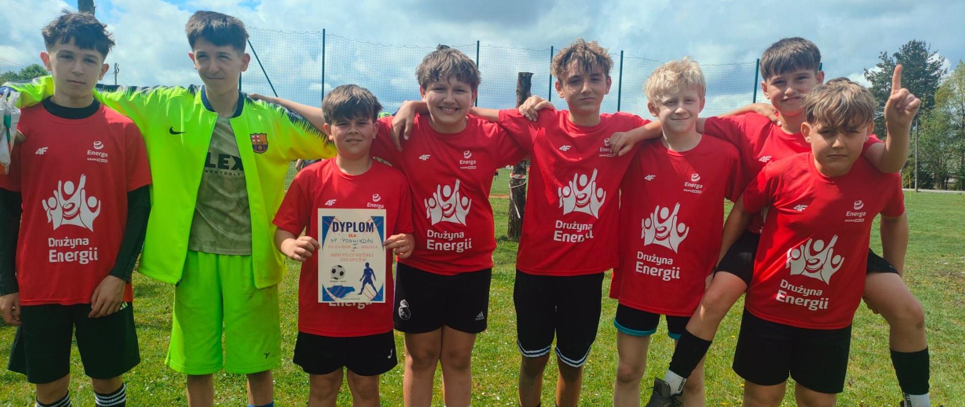 Na zdjęciu widać grupę ośmiu chłopców stojących razem na boisku sportowym. Większość z nich ma na sobie czerwone koszulki sportowe z napisem „Drużyna Energii”, czarne spodenki oraz piłkarskie getry. Jeden z chłopców wyróżnia się jaskrawym, neonowo-zielonym strojem bramkarskim i rękawicami.
Chłopcy stoją ramię w ramię, uśmiechają się i wyglądają na zgraną drużynę po meczu lub turnieju piłkarskim. Jeden z nich trzyma dyplom, co sugeruje, że drużyna zdobyła jakieś wyróżnienie lub miejsce w zawodach. Po prawej stronie jeden z zawodników pokazuje palec uniesiony do góry w geście zwycięstwa lub radości.
W tle widać zielone boisko, siatkę ogrodzeniową, drzewa oraz niebo z dużymi chmurami i przebijającym się słońcem.