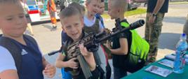 Kilku uczniów stoi przy stoliku żołnierzy, jeden trzyma w ręku karabin, w tle widać karetkę