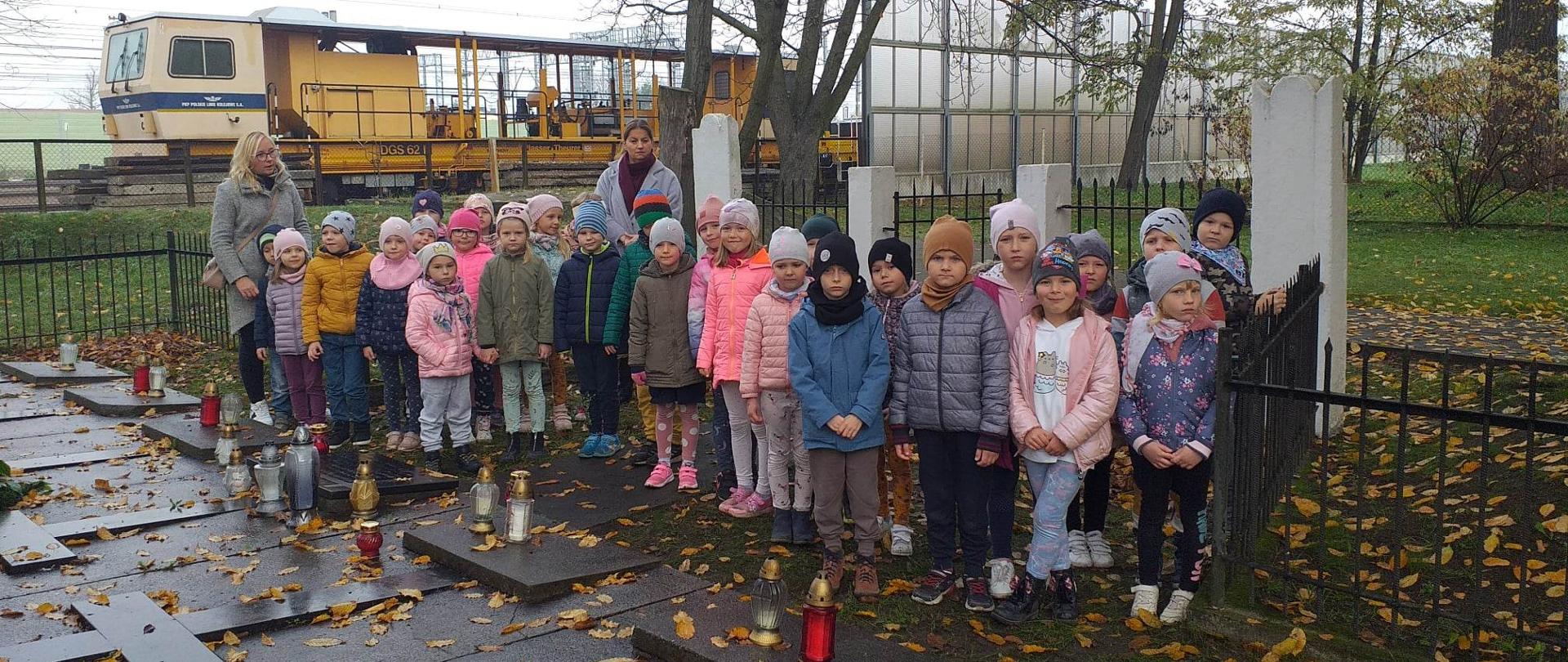 Dzieci stojące przy kamiennych płytach na ziemi. Za nimi stoją dwie kobiety.