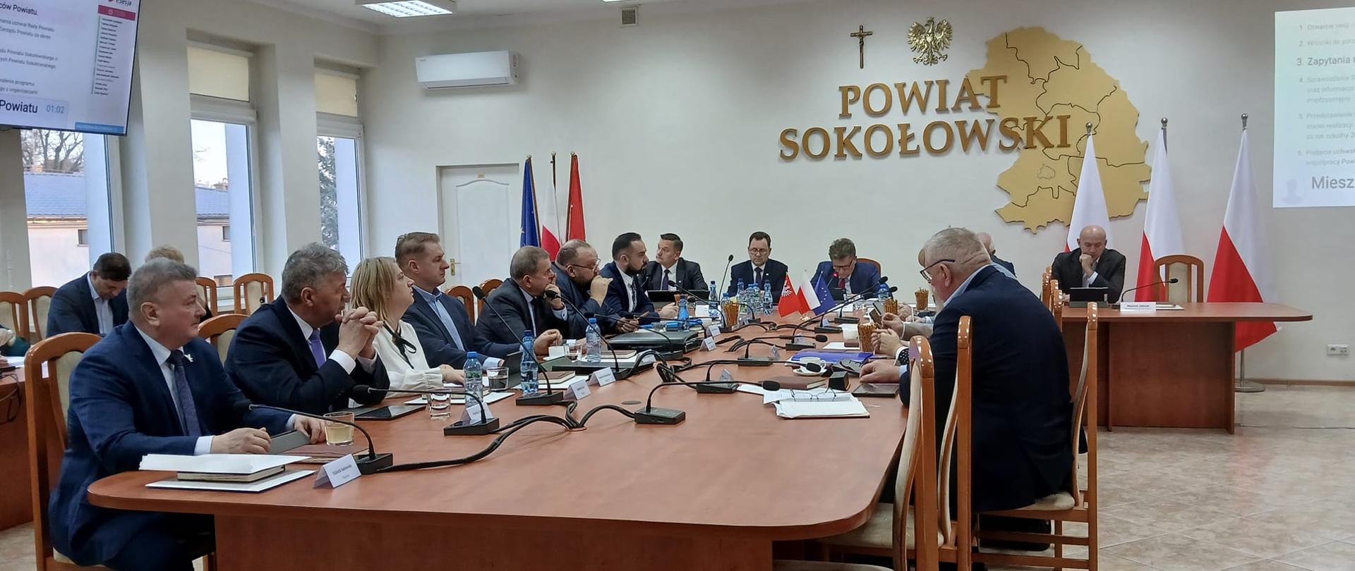 Na zdjęciu widzimy salę konferencyjną Starostwa Powiatowego. W głównym planie długi stół drewniany ustawiony w literę T, przy którym zasiadają radni Rady Powiatu Sokołowskiego. Na przeciwległej ścianie widzimy przestrzenną mapę Powiatu oraz duży napis Powiat Sokołowski. Dodatkowo widzimy flagi biało - czerwone na stojakach oraz flagę Unii Europejskiej i Mazowsza. Nad napisem Powiat Sokołowski wisi krzyż katolicki oraz złotego orła w koronie.