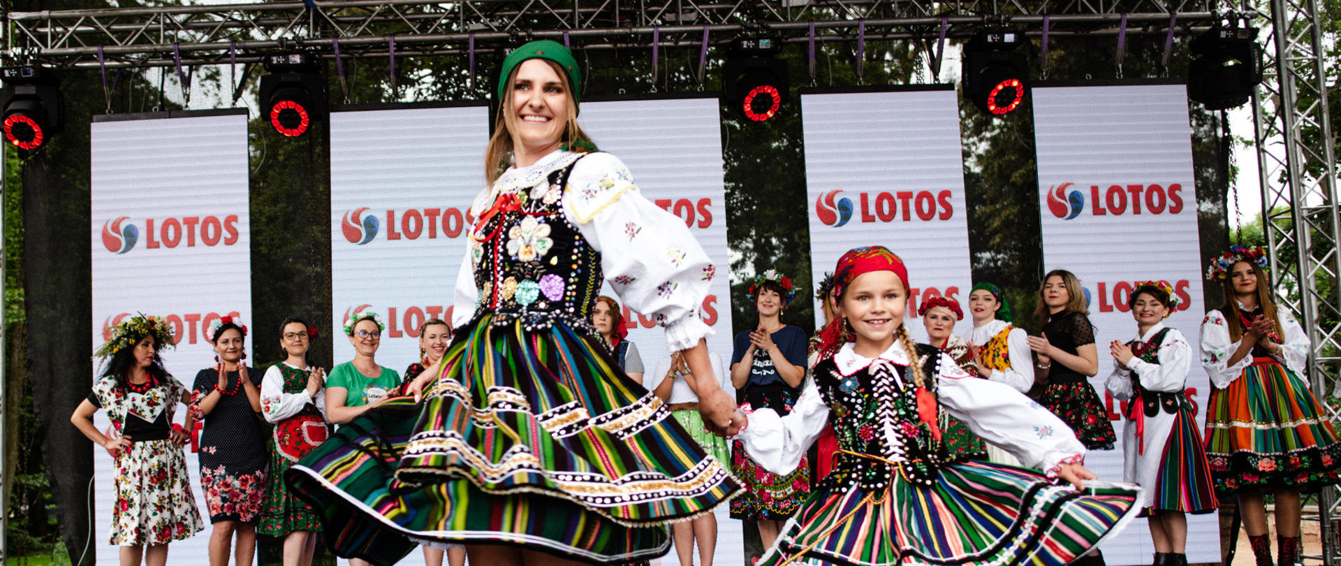 Dorosła kobieta oraz dziewczynka w strojach ludowych tańczą na scenie. W drugim rzędzie stoją w szeregu kobiety w tradycyjnych kolorowych ludowych strojach. Widać również pięć pionowych banerów z logotypem marki Lotos
