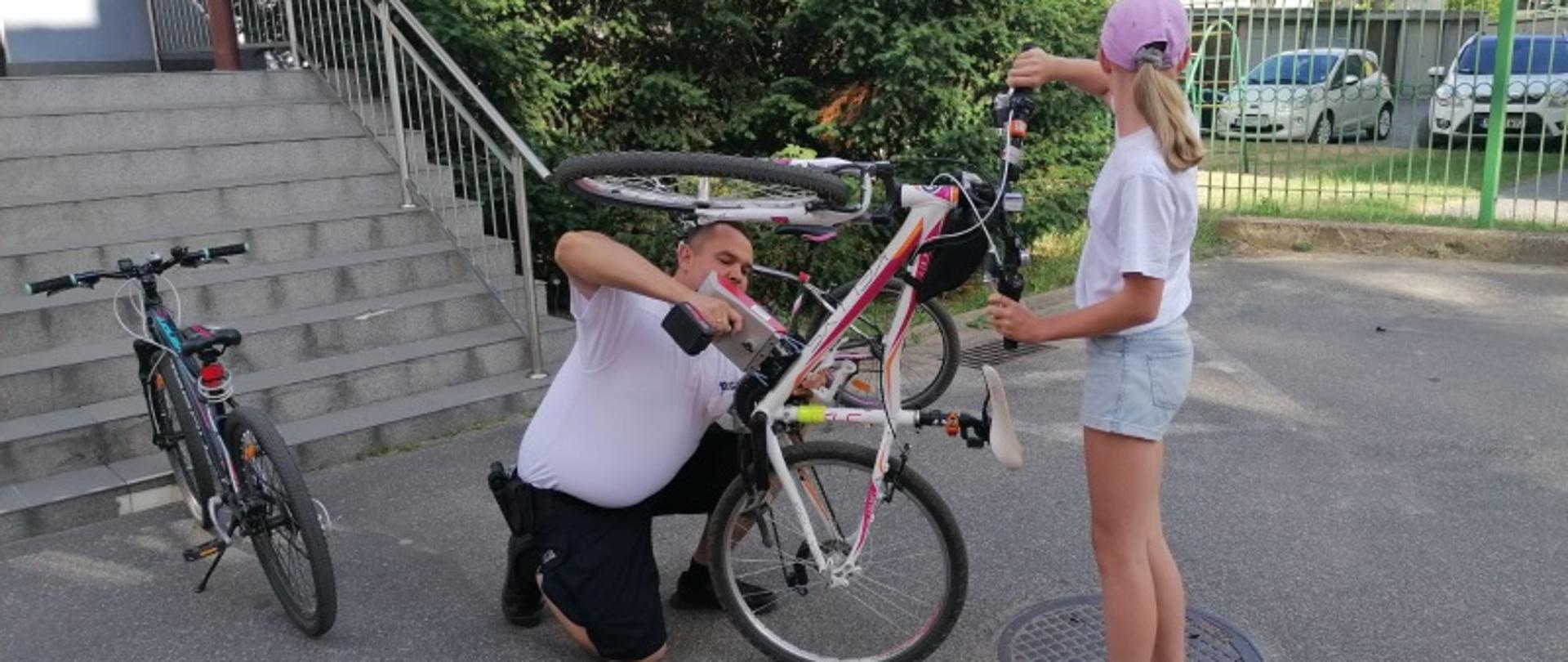 Mężczyzna klęczący przed rowerem, nanoszący specjalnym urządzeniem identyfikator na rower. Rower w pozycji z uniesionym przednim kołem, trzymany przez dziewczynkę