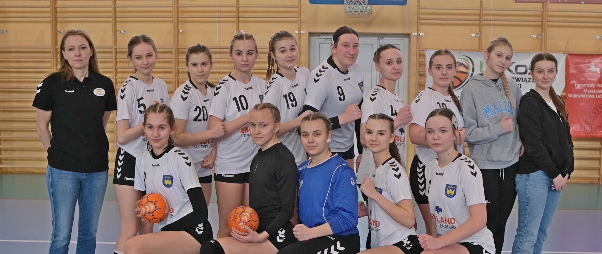 Zdjęcie przedstawia 14 dziewcząt KMKS Kraśnik, stojących na hali sportowej. Pięć zawodniczek klęczy na jedno kolano, zaś kolejne dziewięć stoi ustawione bokiem w tylnym rzędzie. Po lewej stronie zawodniczek stoi trenerka p. Agata Kurlej.
