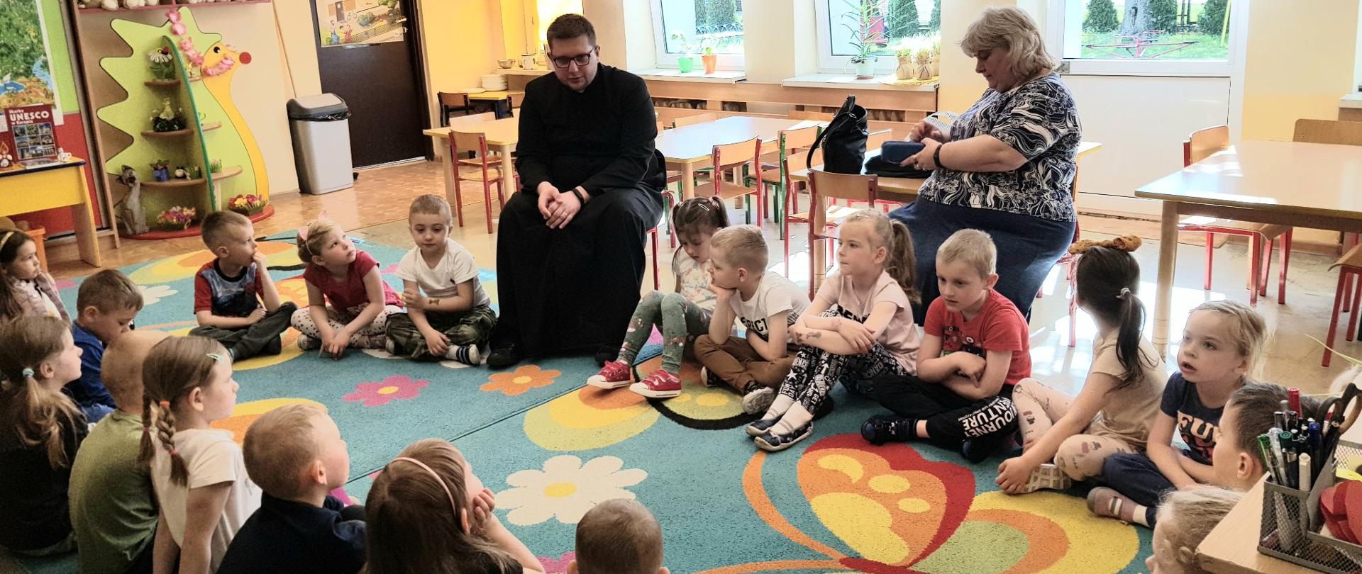 Dzieci siedzące na dywanie z klerykiem Jakubem