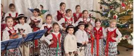Grupa dzieci w strojach góralskich śpiewają i grają kolędy na skrzypcach.