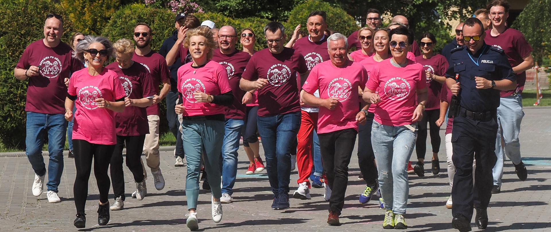 Zdjęcie przedstawia grupę biegnących osób. Uczestnicy biegu ubrani są w różowe koszulki z białym logo "Biegu po uśmiech".