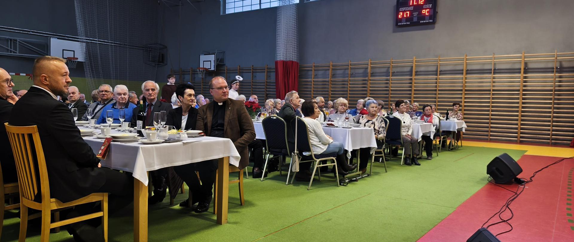 Starsi ludzie siedzą przy stołach w szarej sali gimnastycznej