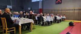 Starsi ludzie siedzą przy stołach w szarej sali gimnastycznej
