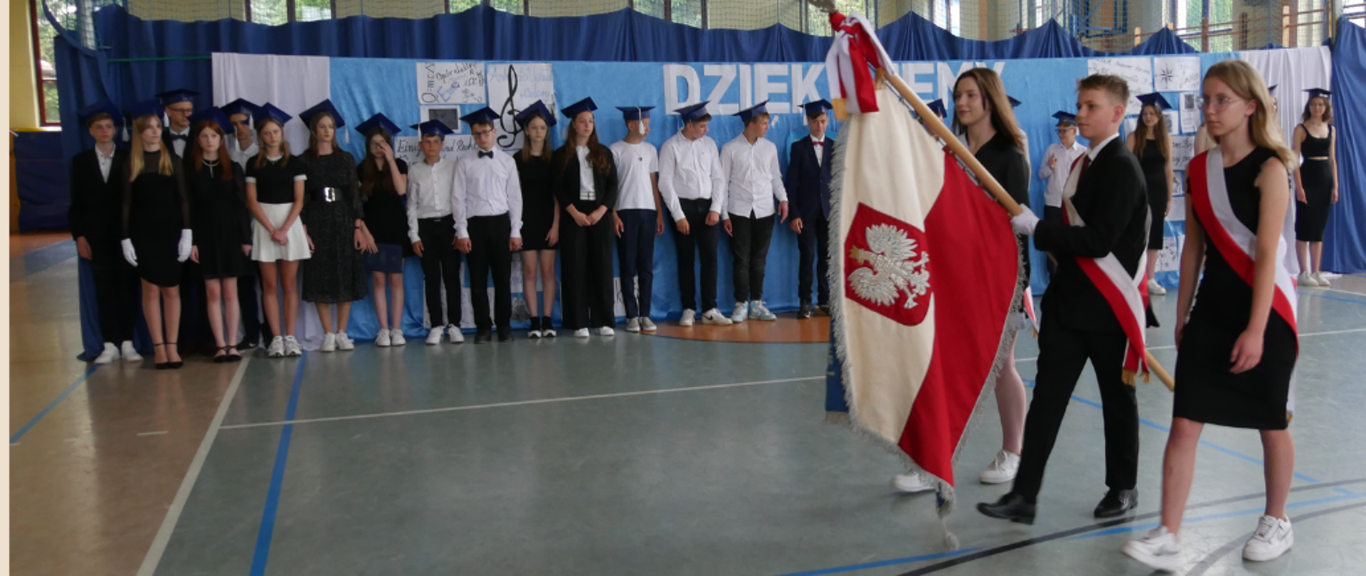 Na hali sportowej uczniowie klasy ósmej ustawieni na niebieskim tle dekoracji z napisem DZIĘKUJEMY, ubrani w galowe stroje w barwach biało- czarno- granatowych. Na głowach mają założone granatowe birety. Po prawej stronie uczniowie ustawieni z pocztem sztandarowym rozpoczynają przemarza.