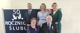 Państwo Krystyna i Kazimierz na tle granatowej tablicy z napisem wraz z Wójtem, Z-cą Wójta i Z-cą Kierownika USC