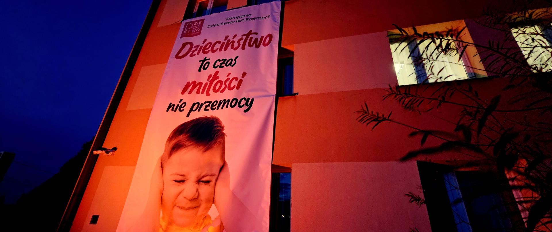 Budynek Poradni Psychologiczno-Pedagogicznej i Powiatowego Centrum Pomocy Rodzinie podświetlony na czerwono. Na jego ścianie widoczny jest baner promujący kampanię „Dzieciństwo bez przemocy”