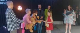 Karolina odbiera gratulacje i nagrodę w postaci dużego brązowego misia od jury konkursu