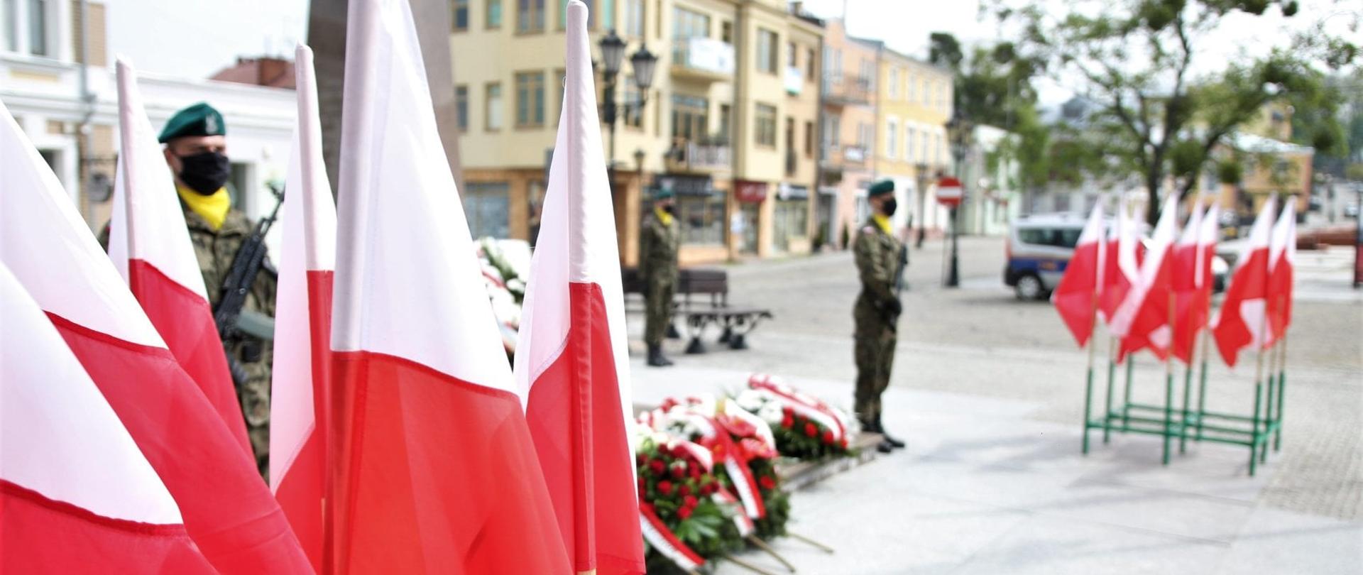 Zdjęcie wykonane na zewnątrz, na placu Łuczkowskiego Chełmie. Na pierwszym tle kilka biało-czerwonych flag. W tle pomnik z położonymi przed nim wieńcami. Na dalszym planie trzej żołnierze z bronią palną.
