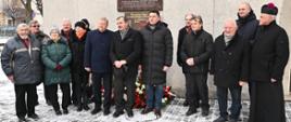 Grupa osób stoi obok siebie przy tablicy "Wdzięczni Bogu za odzyskaną wolność w 25 rocznicę powstania NSZZ". Są ubrani w kurtki, czapki, a dookoła jest śnieg.