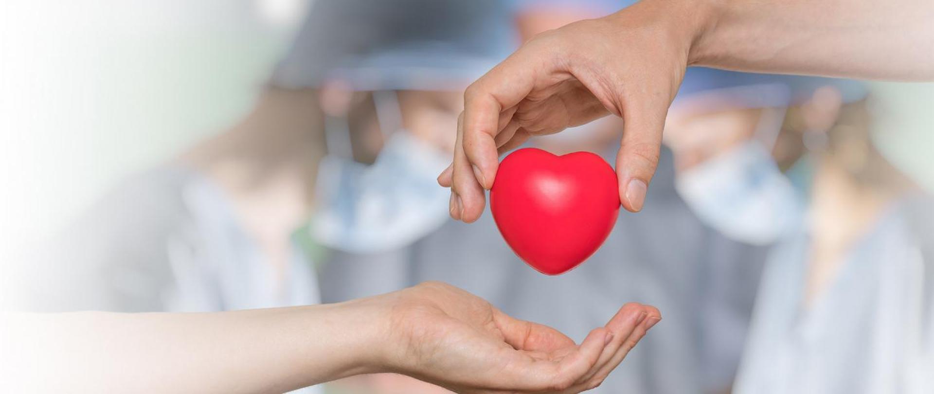 Serce przekazywane z dłoni jednej osoby w dłoń drugiej osoby. W tle lekarze podczas operacji 