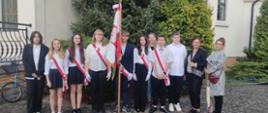 Przed kościołem stoi ośmioro uczniów ubranych w stroje galowe oraz trzy nauczycielki .Po środku stoi chłopak trzymający sztandar szkoły. Sześcioro uczniów ma na koszulach biało-czerwone szarfy. 