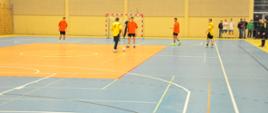 wnętrze sali sportowej w Margoninie, widok na niebiesko-pomarańczowe boisko ze sztuczną nawierzchnią z liniami w kolorach białym, pomarańczowym, żółtym, czarnym, w tle zawodnicy grający w piłkę nożną, za nimi bramka z czerwono-białym obramowaniem