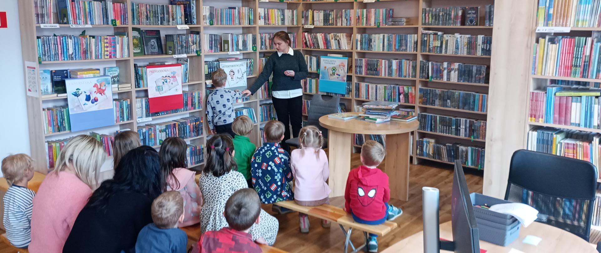 Na zdjęciu widać dzieci w wieku przedszkolnym siedzące na ławeczkach w bibliotece i biorące udział w zabawie przyporządkowywania elementów do odpowiednich gatunków literackich, które przedstawione są na kolorowych plakatach wiszących na regałach z książkami.