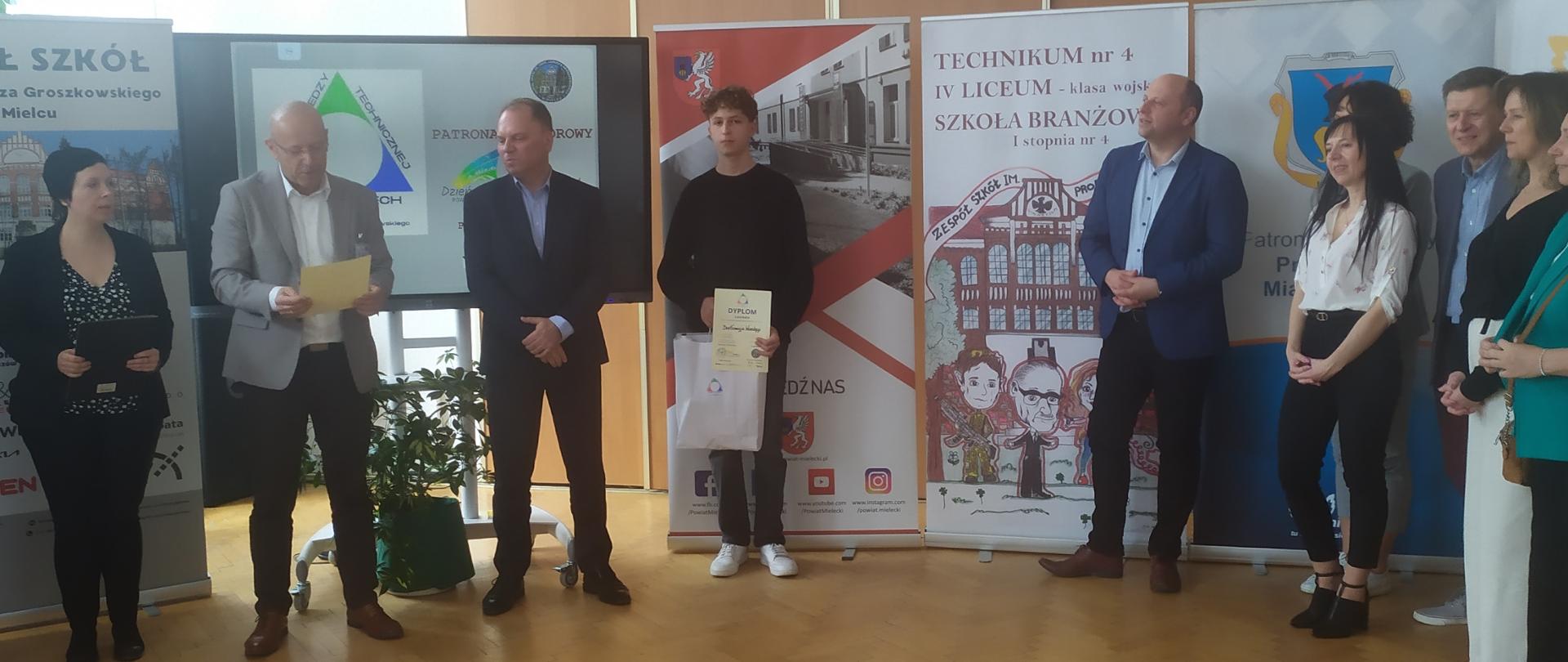 Organizatorzy konkursu, sponsorzy oraz zwycięzca konkursu wraz z dyplomem i nagrodami na tle banerów reklamowych.