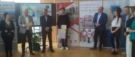 Organizatorzy konkursu, sponsorzy oraz zwycięzca konkursu wraz z dyplomem i nagrodami na tle banerów reklamowych.