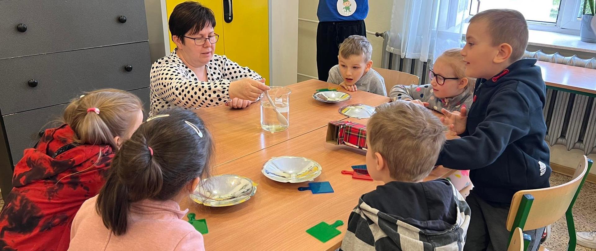 Dzieci siedzą przy ławkach szkolnych oglądają doświadczenia pokazywane przez nauczyciela