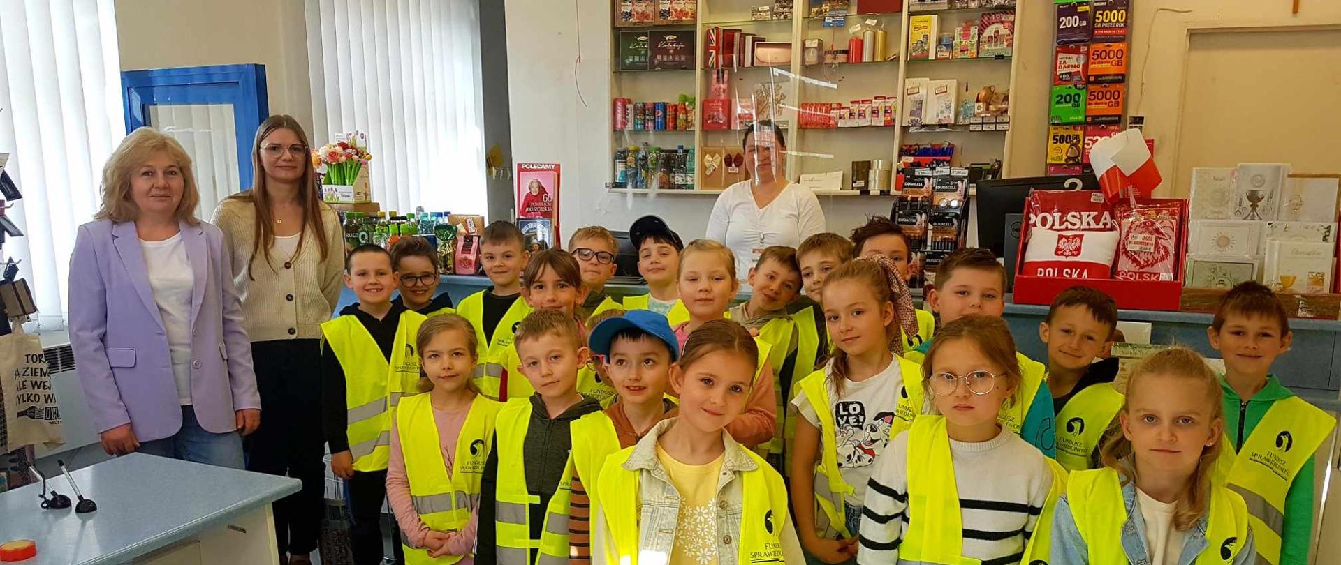 Uczniowie klasy pierwszej w strojach odblaskowych w budynku poczty wraz z Panią wychowawczynią, Panią praktykantką i pracownicą poczty Panią Agnieszką. 
