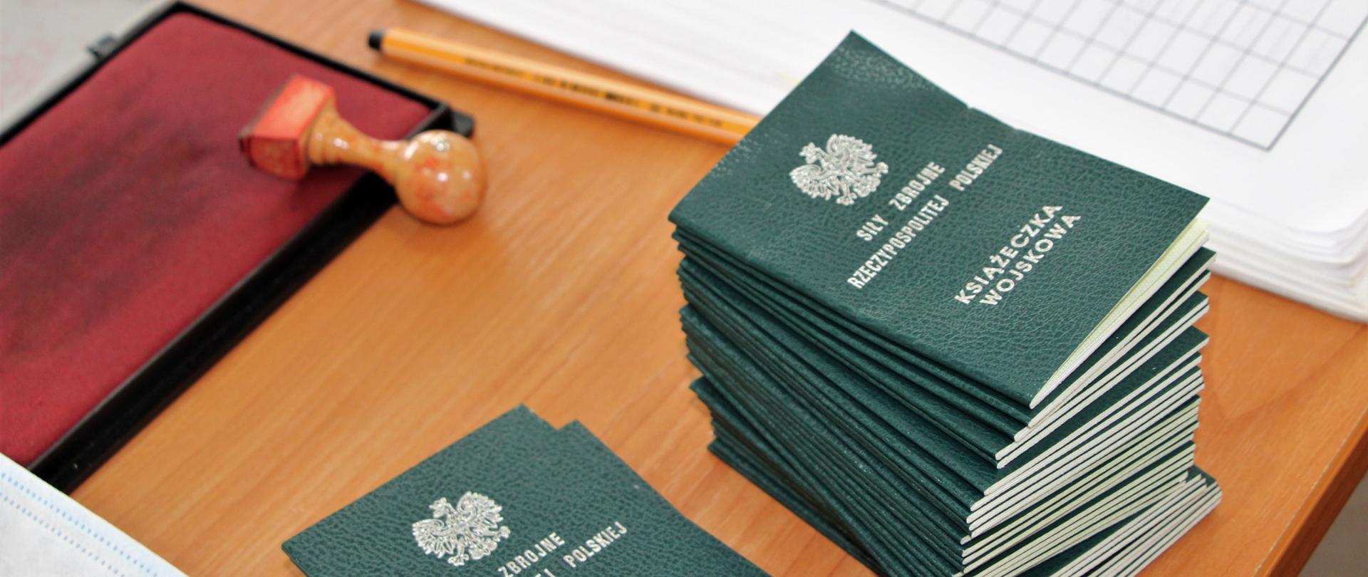 Na biurku leża zielone książeczki wojskowe. Z prawej strony leży maseczka. Obok maseczki leży czerwona pieczątka, długopis i dokumenty.
