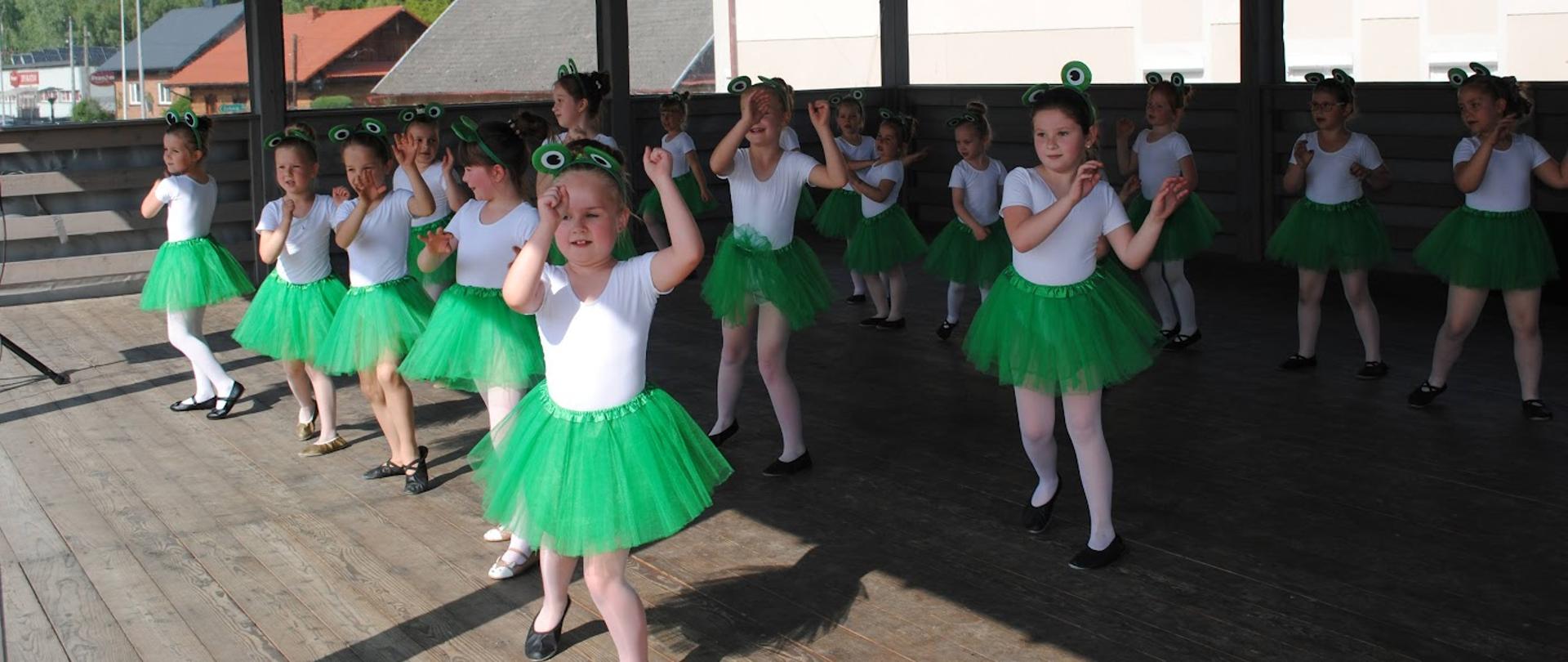 dziewczynki w białych bluzeczkach i zielonych spodniczkach tańczą na drewnianej scenie 