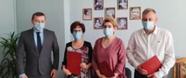 Czwórka ludzi stojących przodem. Dwie kobiety w środku. Troje z nich trzyma w rękach czerwone teczki. Wszyscy mają założone niebieskie maseczki na twarzy. W tle na ścianie wiszą w ramkach obrazki dzieci