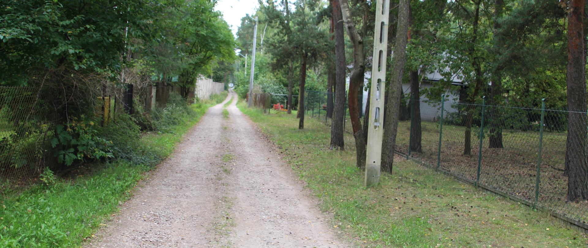Zdjęcie przedstawia polną drogę, która prowadzi w głąb lasu. Droga jest żwirowa, z dwoma widocznymi śladami kół, a po jej obu stronach rosną gęste drzewa. Po prawej stronie widać słup energetyczny oraz siatkę ogrodzeniową.