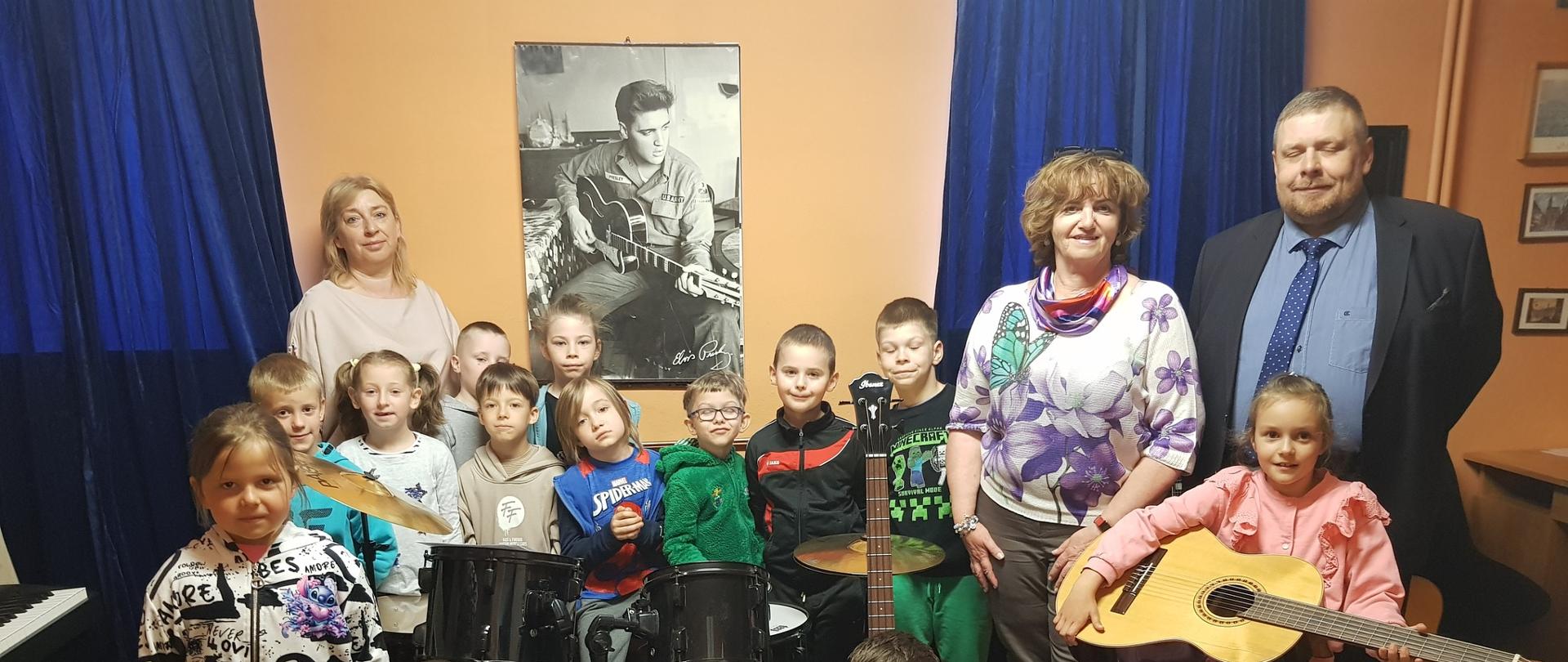 3 dorosłe osoby i grupa dzieci przy instrumentach muzycznych: perkusja, gitara