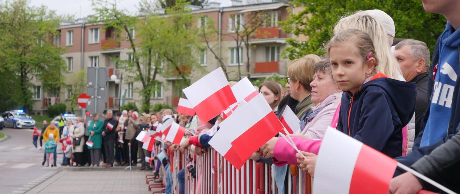 Zdjęcie przedstawia ludzi stojących na zewnątrz, za barierkami i trzymają w rękach biało-czerwone flagi