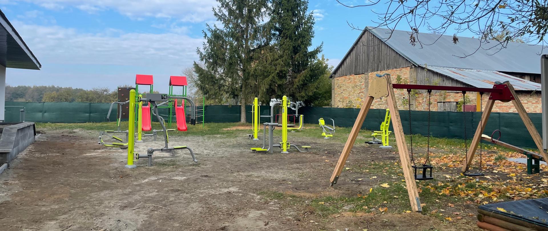 Teren działki z widocznymi urządzeniami siłowni zewnętrznej i placu zabaw. Za urządzeniami widoczne dwa duże świerki. W tle błękitne niebo z chmurami. Z prawej strony widoczny budynek gospodarczy. W prawym górnym rogu widoczna gałąź drzewa. 