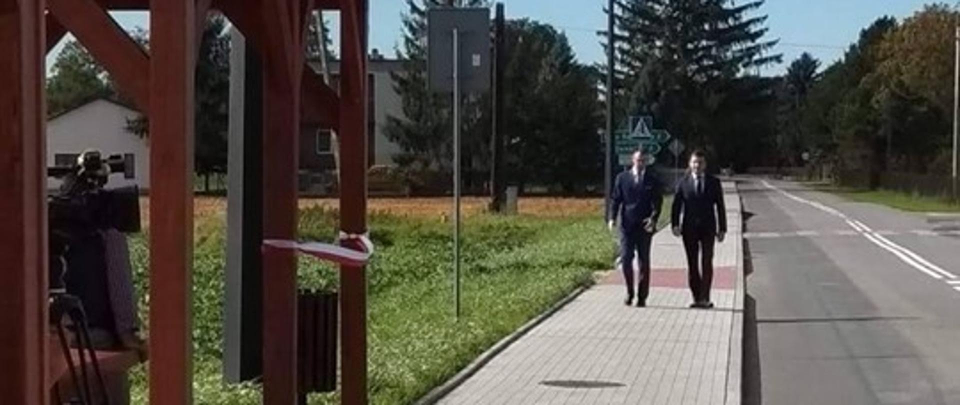 zdjęcie przedstawia fragment ścieżki rowerowej wykonanej z kostki brukowej w kolorze czerwonym i szarym. Z lewej strony widać fragment wiaty przystankowej. W tle Wójt Gminy Wietrzychowice oraz Przewodniczący Komisji Budżetu i Radny Województwa Małopolskiego