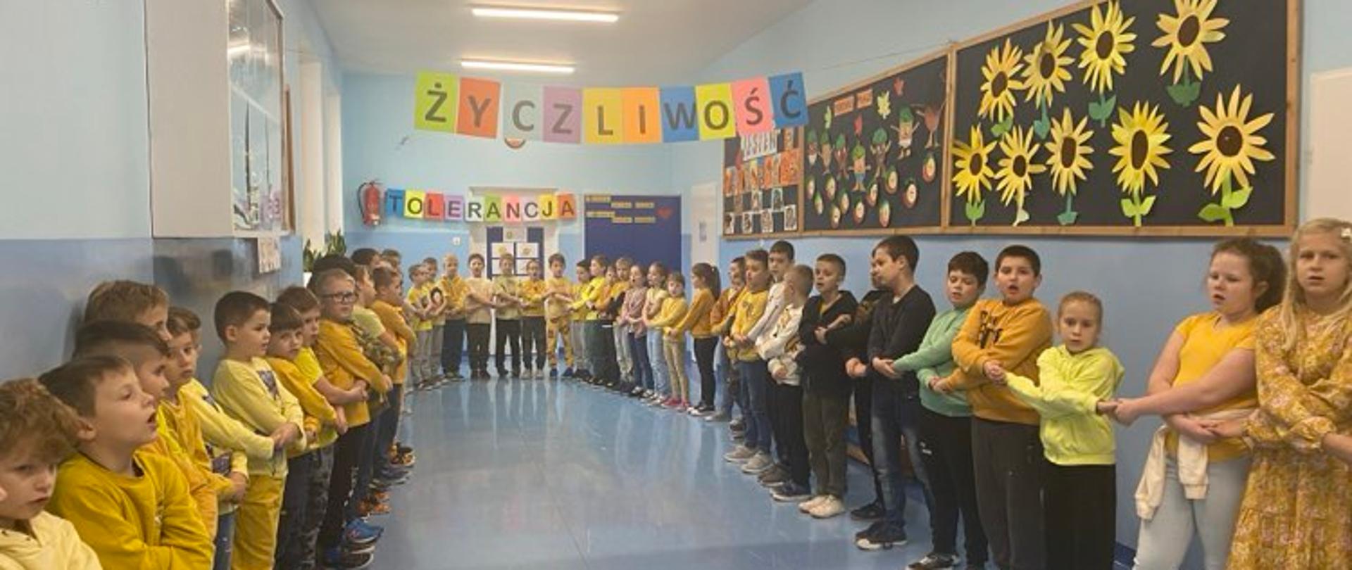 Na szkolnym korytarzu stoją uczniowie ubrani na żółto, trzymają się za ręce i tworzą krąg. U góry rozwieszone napisy: życzliwość, tolerancja. Z prawej strony na gazetce są żółte słoneczniki.
