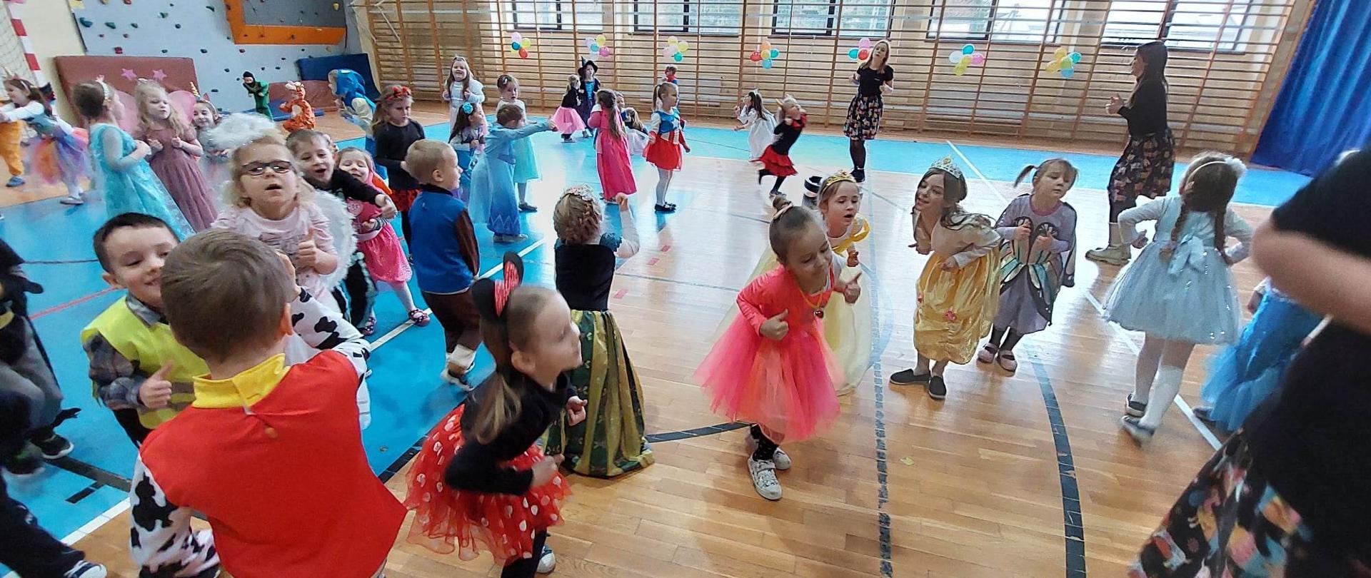 Radosne i uśmiechnięte dzieci w kolorowych, bajkowych kostiumach tańczą podczas balu karnawałowego na sali gimnastycznej. 
