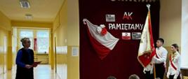 Pani dyrektor przemawia do uczniów, po prawej stronie stoi poczet sztandarowy szkoły ze sztandarem, za nimi bordowa zasłona, na której przypięto biało czerwoną flagę, napis "AK pamiętamy" oraz symbol Polski walczącej, na pierwszym planie widać uczniów oglądających akademię