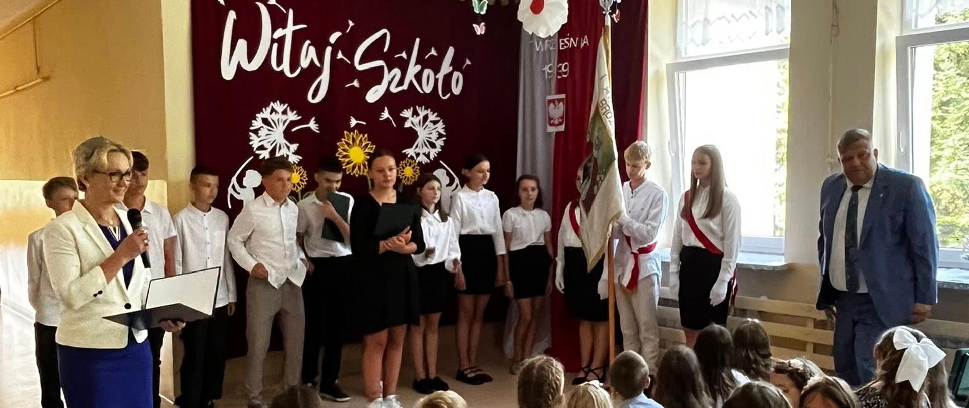 Pani dyrektor przemawia do mikrofonu, po prawej stronie uczniowie ubrani na galowo prezentują występ artystyczny z okazji rozpoczęcia roku szkolnego, obok nich stoi poczet sztandarowy i wójt Gminy Łabowa, za nimi na ścianie na bordowym płótnie napis "Witaj szkoło" oraz wycięte z białego kartonu dmuchawce, przed występującymi na ławeczkach siedzą dzieci
