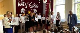 Pani dyrektor przemawia do mikrofonu, po prawej stronie uczniowie ubrani na galowo prezentują występ artystyczny z okazji rozpoczęcia roku szkolnego, obok nich stoi poczet sztandarowy i wójt Gminy Łabowa, za nimi na ścianie na bordowym płótnie napis "Witaj szkoło" oraz wycięte z białego kartonu dmuchawce, przed występującymi na ławeczkach siedzą dzieci