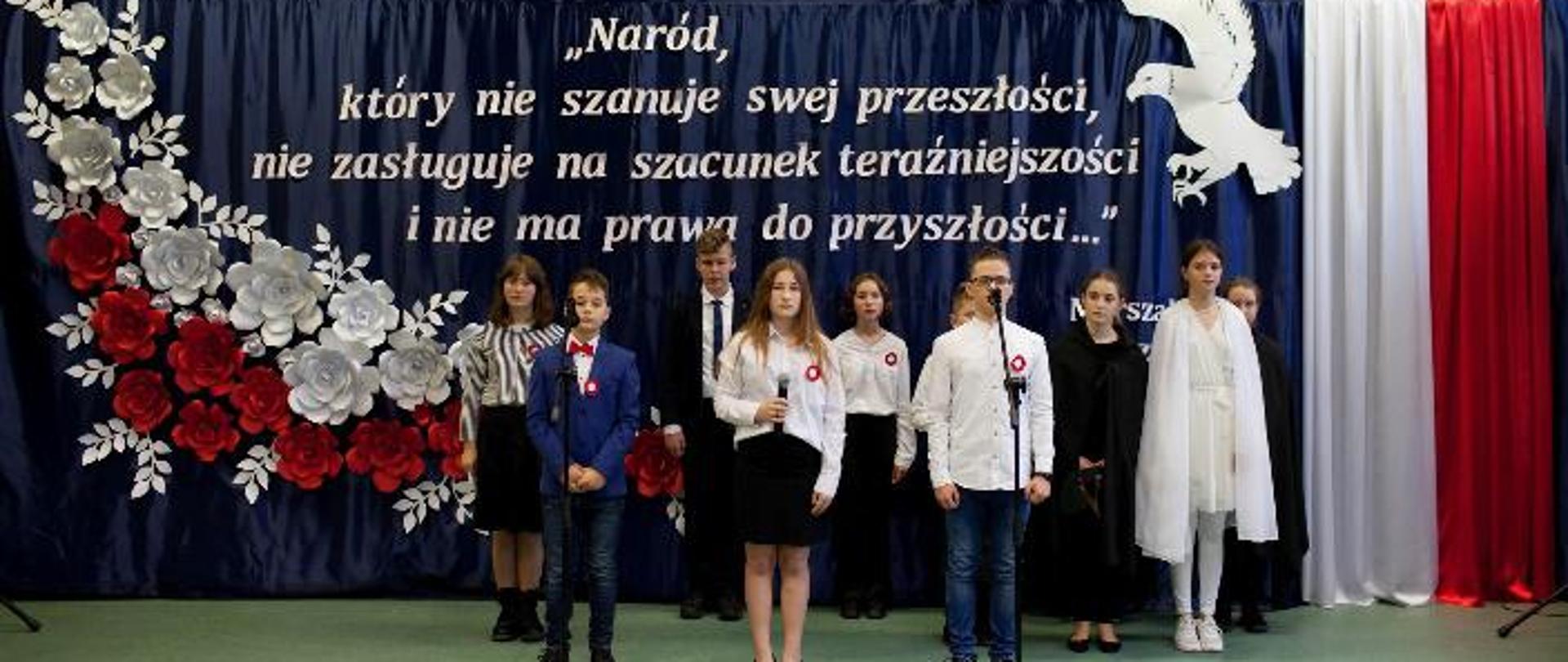 Zdjęcie przedstawia uczniów uczestniczących w akademii szkolnej. Uczniowie na tle sceny z biało-czerwonych kwiatów i cytatu J. Piłsudskiego oraz białego orła