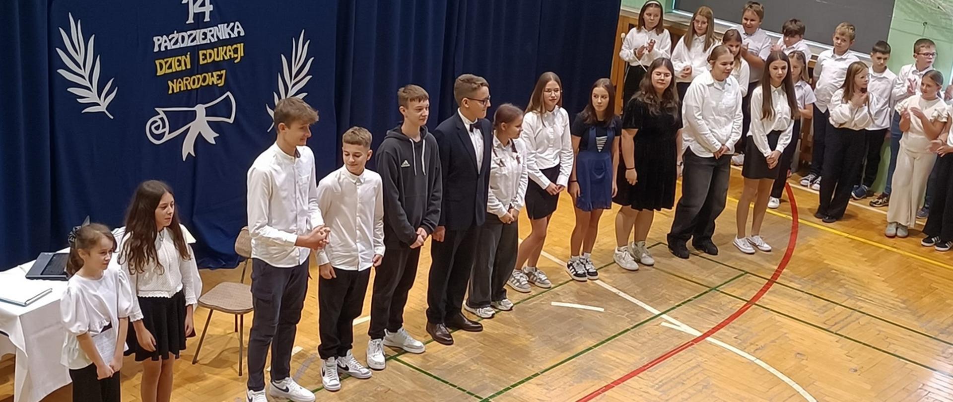 Grupa dzieci i młodzieży ubrana na galowo stoi w szeregu w szkolnej sali gimnastycznej. Za nimi dekoracja z napisem: 14 października Dzień Edukacji Narodowej.