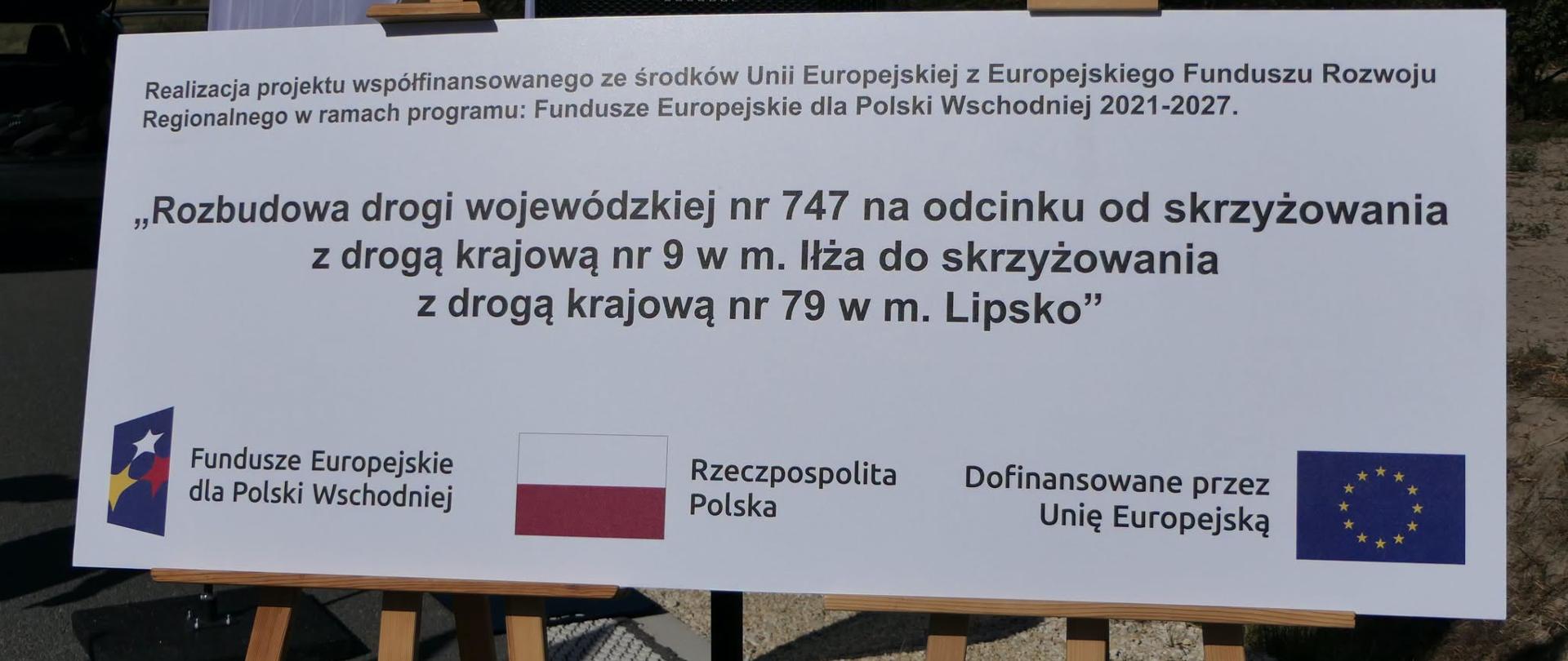 Biała tablica informacyjna na drewnianych sztalugach z nazwą projektu rozbudowy drogi nr 747 i logotypami unijnymi.