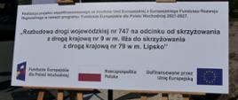 Biała tablica informacyjna na drewnianych sztalugach z nazwą projektu rozbudowy drogi nr 747 i logotypami unijnymi.