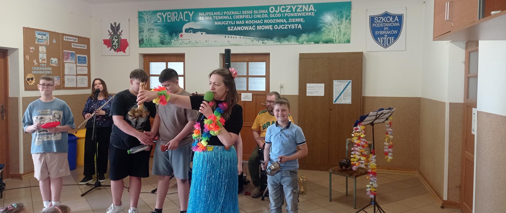 Na przodzie stoi kobieta w niebieskiej połyskującej spódnicy. Na szyi ma kolorową girlandę. Za nią stoi czworo uczniów. Każdy z nich trzyma w dłoni jakiś przedmiot - miskę, butelkę, kołatkę bądź garnek