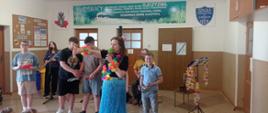 Na przodzie stoi kobieta w niebieskiej połyskującej spódnicy. Na szyi ma kolorową girlandę. Za nią stoi czworo uczniów. Każdy z nich trzyma w dłoni jakiś przedmiot - miskę, butelkę, kołatkę bądź garnek
