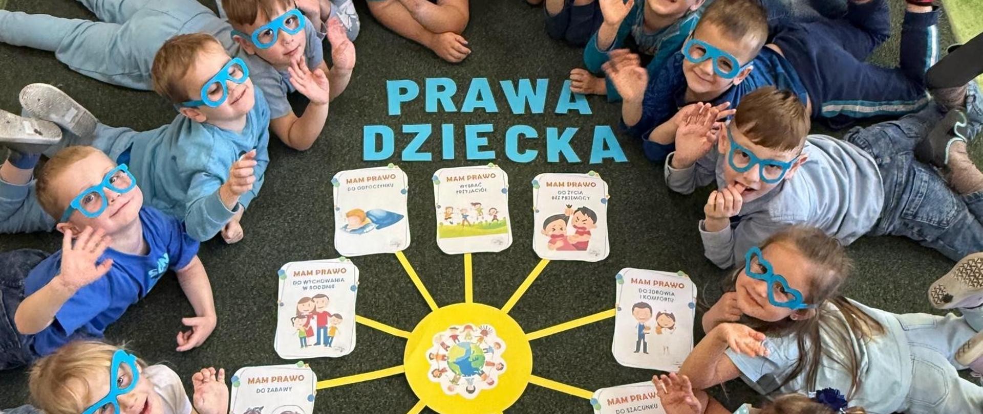 Przedszkolaki leżą na brzuchu na dywanie, podnoszą głowy do góry, ubrani są na niebiesko, na nosie mają niebeskie okulary, pomiędzy nimi na dywanie ułożony z niebieskich liter napis "Prawa dziecka" oraz wycięte z papieru żółte słoneczko, na końcach jego promieni znajdują się ilustracje z prawami dziecka