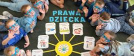 Przedszkolaki leżą na brzuchu na dywanie, podnoszą głowy do góry, ubrani są na niebiesko, na nosie mają niebeskie okulary, pomiędzy nimi na dywanie ułożony z niebieskich liter napis "Prawa dziecka" oraz wycięte z papieru żółte słoneczko, na końcach jego promieni znajdują się ilustracje z prawami dziecka